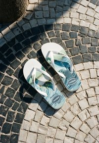 Tongs blanches Havaianas avec un motif de vagues océaniques bleues reposant sur un sol en mosaïque noir et blanc près d'un tronc d'arbre.