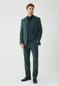 Uomo che indossa un completo tre pezzi verde scuro con camicia nera e cravatta, in piedi con una mano in tasca su sfondo bianco semplice.