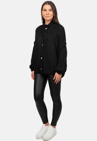 Zwarte gebreide cardigan met een hoge kraag en grote knopen, gedragen over glanzende zwarte leggings en witte sneakers.