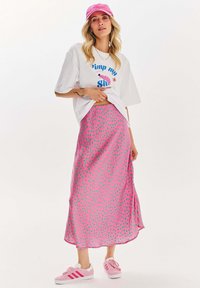 Růžová saténová maxi sukně s zelenými puntíky, doplněná bílým tričkem s grafikou a růžovou kšiltovkou. Bílé tenisky dotvářejí celkový vzhled.