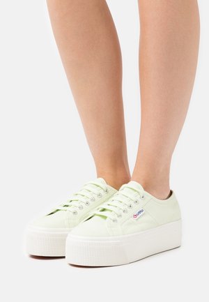 Jambes portant des baskets compensées en toile vert clair avec des semelles épaisses en caoutchouc blanc et des lacets, debout sur un fond blanc.