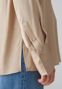 Beige Langarmshirt mit glatter Textur, ausgestattet mit geknöpften Manschetten und seitlichen Schlitzen, kombiniert mit blauer Denim.