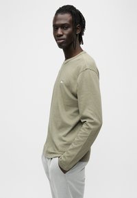 Μακριά μπλούζα σε ελιά πράσινο χρώμα από μαλακό ύφασμα με άνετη εφαρμογή και στρογγυλή λαιμόκοψη, με μικρή λεπτομέρεια λογότυπου στο στήθος.