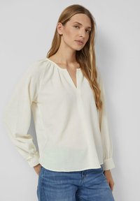 Blusa bianca in crepe con scollatura a V, maniche lunghe a sbuffo e dettagli arricciati sulle spalle. Tessuto con un aspetto testurizzato.