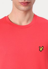 Röd bomulls T-shirt med rundad halsringning, ribbad textur, som har en liten svart och gul örnlogotyp på vänster bröst.