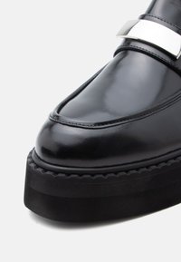 Μαύρα δερμάτινα loafers με γυαλιστερό φινίρισμα, που διαθέτουν εντυπωσιακή ασημένια αγκράφα και χοντρή μαύρη υφασμάτινη σόλα.