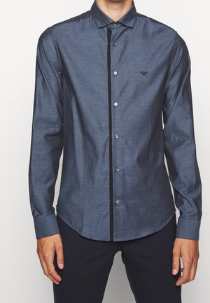 Mężczyzna ubrany w dopasowaną, długą, ciemnoniebieską koszulę na guziki z małym logo na lewej piersi oraz czarne spodnie.