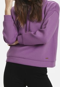 Sweat à capuche violet en tissu doux, avec une coupe décontractée, des manches resserrées aux poignets et un petit logo doré sur l'ourlet.