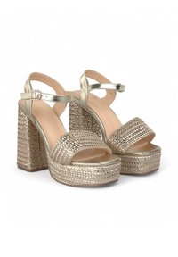 Talons compensés à bout ouvert avec une base tissée texturée dorée, des brides en or métallique et un design de talon épais. Fermeture par boucle sur la bride de cheville.