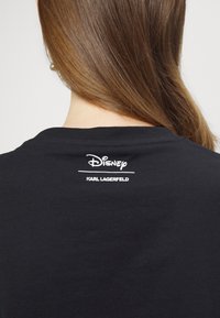 Černé bavlněné tričko s bílým potiskem na zádech: „Disney“ nad „KARL LAGERFELD“. Klasický kulatý výstřih a krátké rukávy.