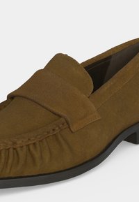 Mocassin en daim marron avec une semelle noire, bout arrondi et détails cousus sur l'avant et les côtés.