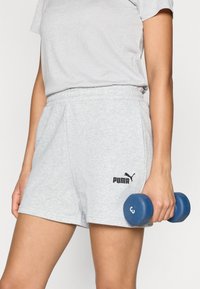 Γκρι αθλητικό σορτς με ελαστική μέση και λογότυπο PUMA, συνδυασμένο με ανοιχτό γκρι μπλουζάκι. Ένα μπλε βαράκι κρατιέται στο δεξί χέρι.