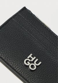Portefeuille noir texturé avec plusieurs emplacements pour cartes, arborant un logo argenté portant l'inscription "HUGO". Forme compacte et rectangulaire.