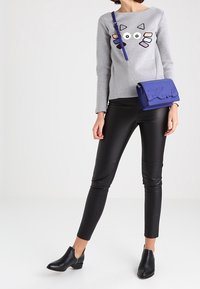 Žena v šedém svetru s barevným potiskem kočičí tváře, černých kožených kalhotách, černých kotníkových botách a modré crossbody tašce.