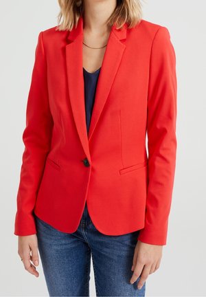 Frau trägt einen roten Blazer über einem dunklen Oberteil mit blauen Jeans vor einem einfarbigen hellen Hintergrund.