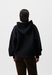 Felpa nera oversize con un grande cappuccio, polsini a coste e una texture morbida. Indossata con pantaloni in velluto a coste marroni. Design semplice e casual.