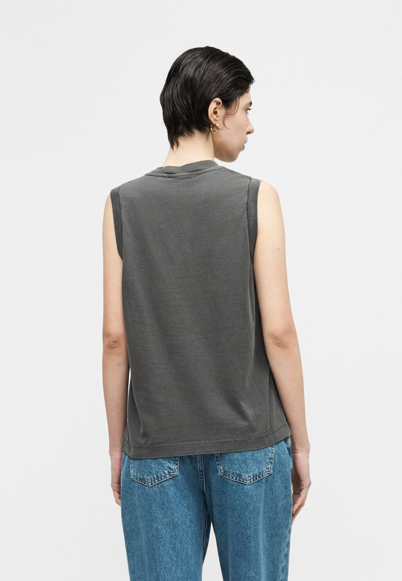 Person with short dark hair, wearing sleeveless dark gray top and blue jeans, facing away against a plain light background.