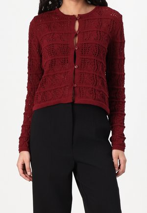 Femme portant un cardigan en tricot bordeaux à manches longues avec des boutons et un pantalon taille haute noir, sur un fond uni.