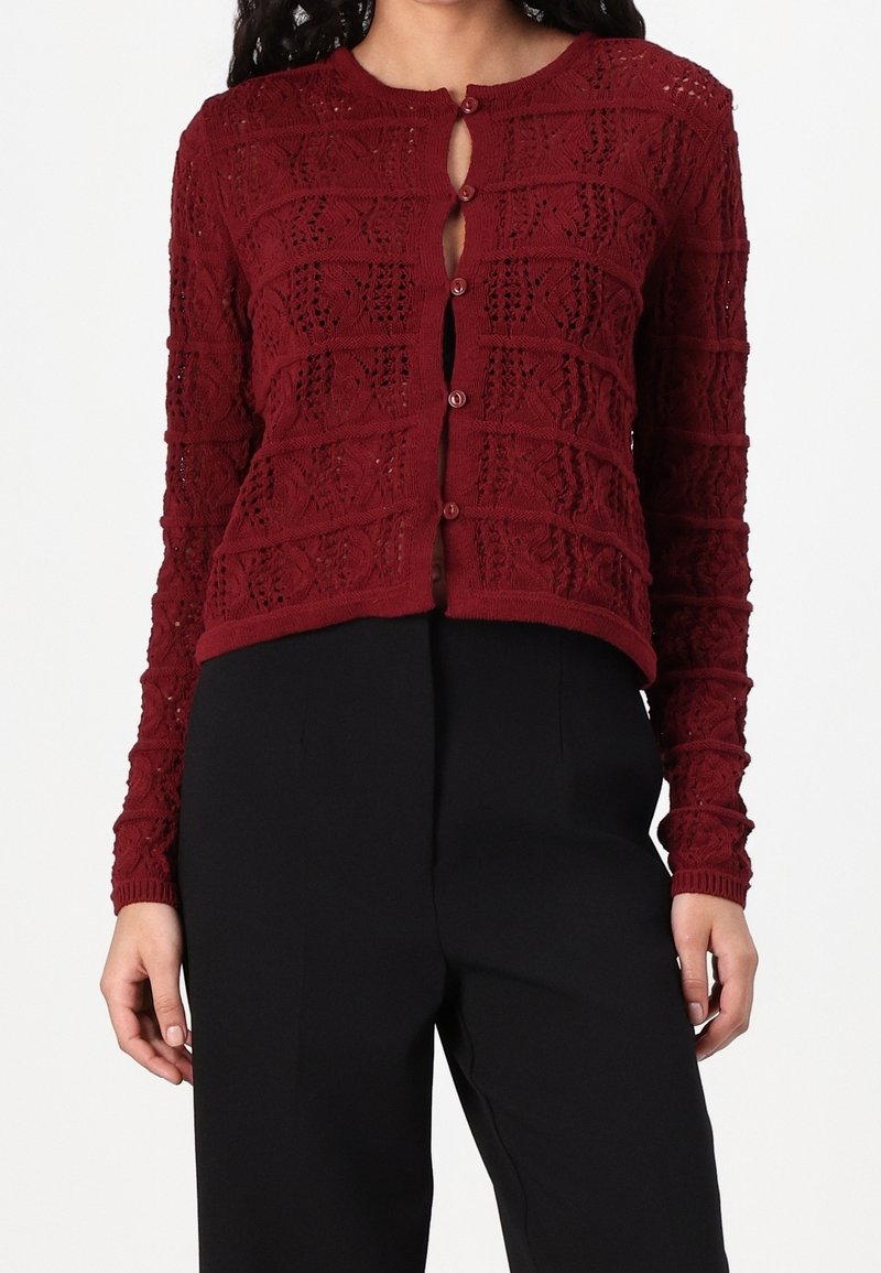 Femme portant un cardigan en tricot bordeaux à manches longues avec des boutons et un pantalon taille haute noir, sur un fond uni.
