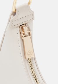 Gros plan sur la fermeture éclair d'un sac en cuir beige avec tirette en métal doré et détails de coutures sur une surface lisse.