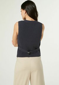 Mornarska pletena bezrukavna majica s teksturiranim uzorkom i detaljem od tkanine u pojasu, u kombinaciji s bež hlačama.