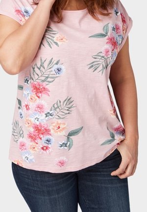 Frau trägt ein hellrosa kurzärmeliges Hemd mit mehrfarbigen Blumenmuster und dunkelblaue Jeans, eine Hand an der Hüfte, die andere in der Nähe des Gesichts.