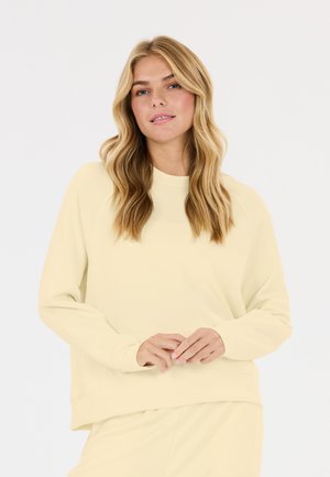 Blonde vrouw met golvend haar, die een lichtgele top met lange mouwen en bijpassende broek draagt, staande voor een effen witte achtergrond.