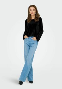 Top a maniche lunghe in velluto nero abbinato a jeans larghi blu chiaro e stivaletti neri. Il modello è in piedi con le mani nelle tasche, rivolgendosi in avanti.