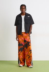 Mand står på grønne gulv mod hvid væg, iført sort kortærmet skjorte over hvid tanktop, brede orange mønstrede bukser og hvide sneakers.