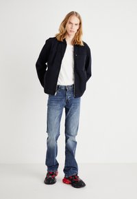 Schwarze Jacke mit Reißverschluss über einem weißen T-Shirt, kombiniert mit blauen Jeans und schwarzen/roten Sneakers. Das Outfit bietet einen lässigen, geschichteten Look.