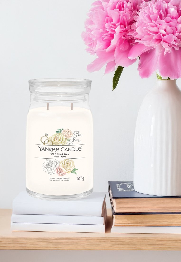 Yankee Candle in einem klaren Glasbehälter mit einem weißen Wachskörper, etikettiert mit "Hochzeitstag", mit floralen Illustrationen, steht auf gestapelten Büchern.