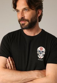 Zwart katoenen t-shirt met een grafische print van een schedel met decoratieve elementen, met de tekst "Deeluxe" onder het ontwerp.