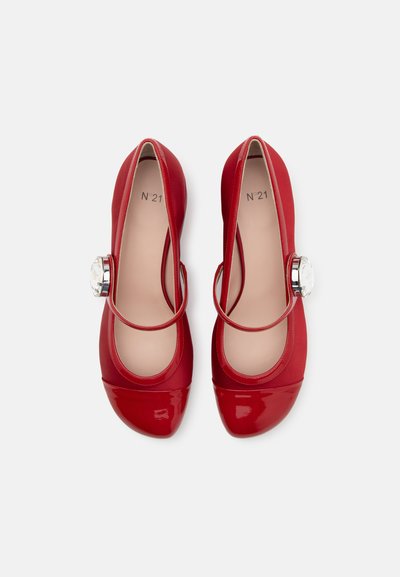 Piros lakkbőr balettcipők fényes orrrésszel, elasztikus pánttal és egy átlátszó drágakő díszítéssel az oldalon. Az anyag sima textúrájú.
