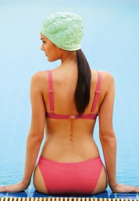 Rosa Badebekleidung mit dünnen Trägern, kombiniert mit einer strukturierten mintgrünen Badekappe mit floralen Mustern. Sitzt am Pool und zeigt glatte Haut.