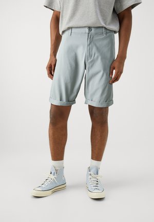 Pantalones cortos de algodón de color azul claro con dobladillo doblado, frente plano y cierre de botón, combinados con zapatillas altas de color azul claro y calcetines blancos.