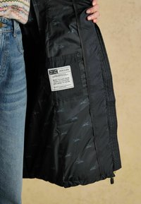 Melns pūkains jaciņš ar quiltotu dizainu, ar rakstainu iekšpusi un etiķeti, kas ir iešūta oderē. Redzama auduma tekstūra un detaļas.