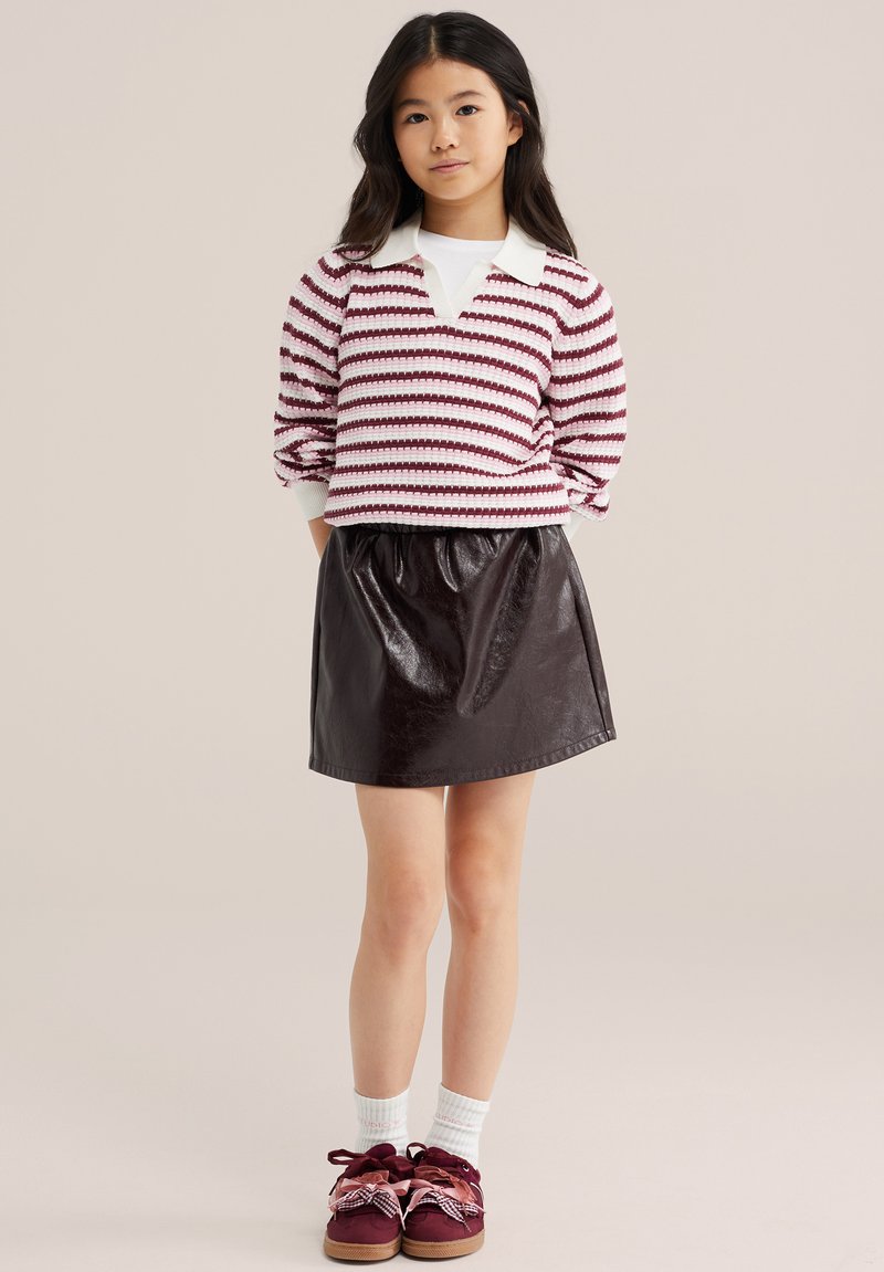 Jeune fille debout, les mains dans le dos, portant un pull rayé bordeaux et blanc, une jupe noire, des chaussettes blanches et des chaussures bordeaux avec des nœuds.