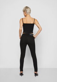 Rövid szőke hajú személy fekete spagettipántos felsőt, fekete skinny farmert és fekete magassarkú szandált visel, keze a csípőjén áll.