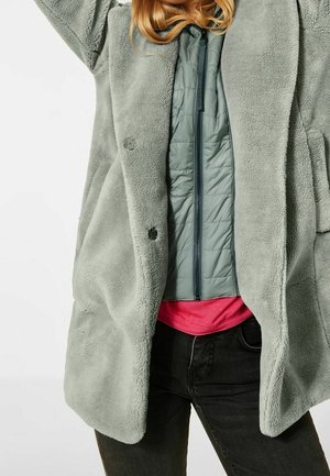 E fluffy, lichtgroene fleecejas met knopen, gelaagd over een donkere geïsoleerde jas en een roze shirt; zwarte jeans zichtbaar bij de taille.