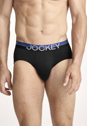 Männlicher Oberkörper, der schwarze Slips mit einem blauen Bund trägt, auf dem "JOCKEY" steht, vor weißem Hintergrund.