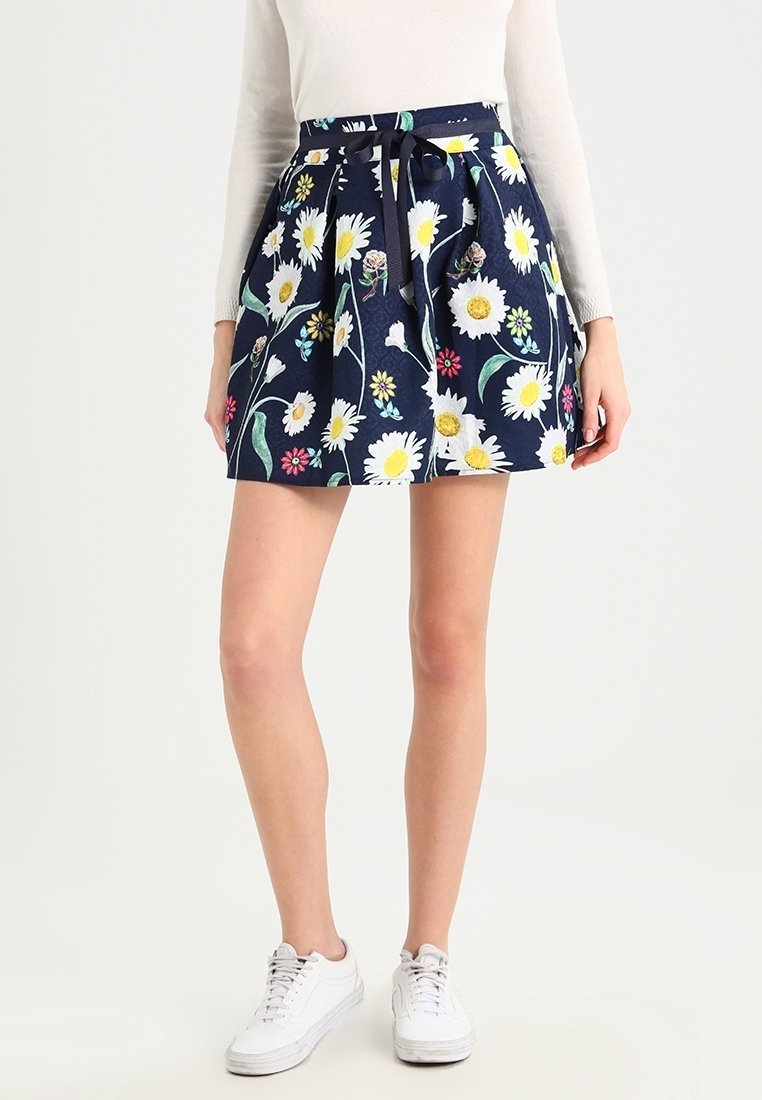Femme portant une jupe bleu marine avec des imprimés de marguerites blanches et jaunes, taille nouée, haut blanc à manches longues et baskets blanches, debout devant un fond uni.