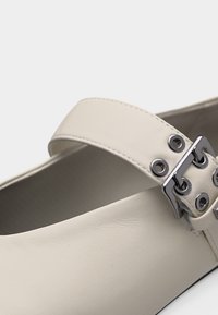 Κοντινό πλάνο λευκού δερμάτινου λουριού παπουτσιού με ασημένια αγκράφα και μεταλλικά τρύπια σε λεία, καμπυλωτή επιφάνεια.