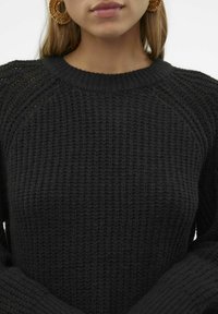 Pull en tricot noir avec un motif texturé, un col et des poignets côtelés, aux manches larges, associé à de grandes boucles d'oreilles circulaires.