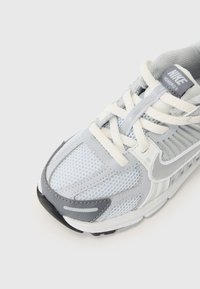 Svijetlosivi atletski tenisice napravljene od mrežastog i sintetičkog materijala, s teksturiranim dizajnom, bijelim vezicama i crnim gumenenim potplatom. Nike logo na jeziku.