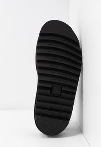 Semelle de chaussure en caoutchouc noir avec des rainures horizontales profondes et un talon texturé, positionnée verticalement contre un mur d’angle blanc.
