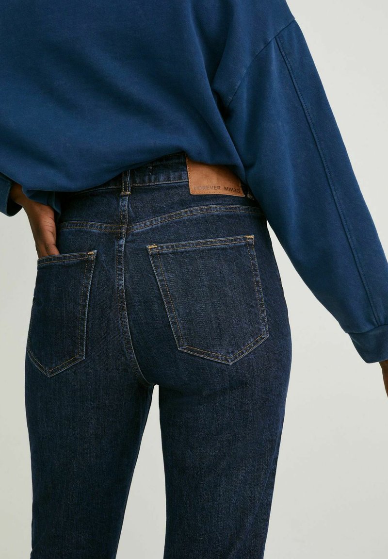 Jean en denim bleu foncé avec une taille haute, offrant deux poches arrière et une étiquette en cuir brun à la taille. Texture lisse et coupe ajustée.
