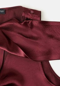 Top de satén burdeos con una textura suave, que presenta un detalle de botón en el cuello y un diseño de hombros descubiertos.