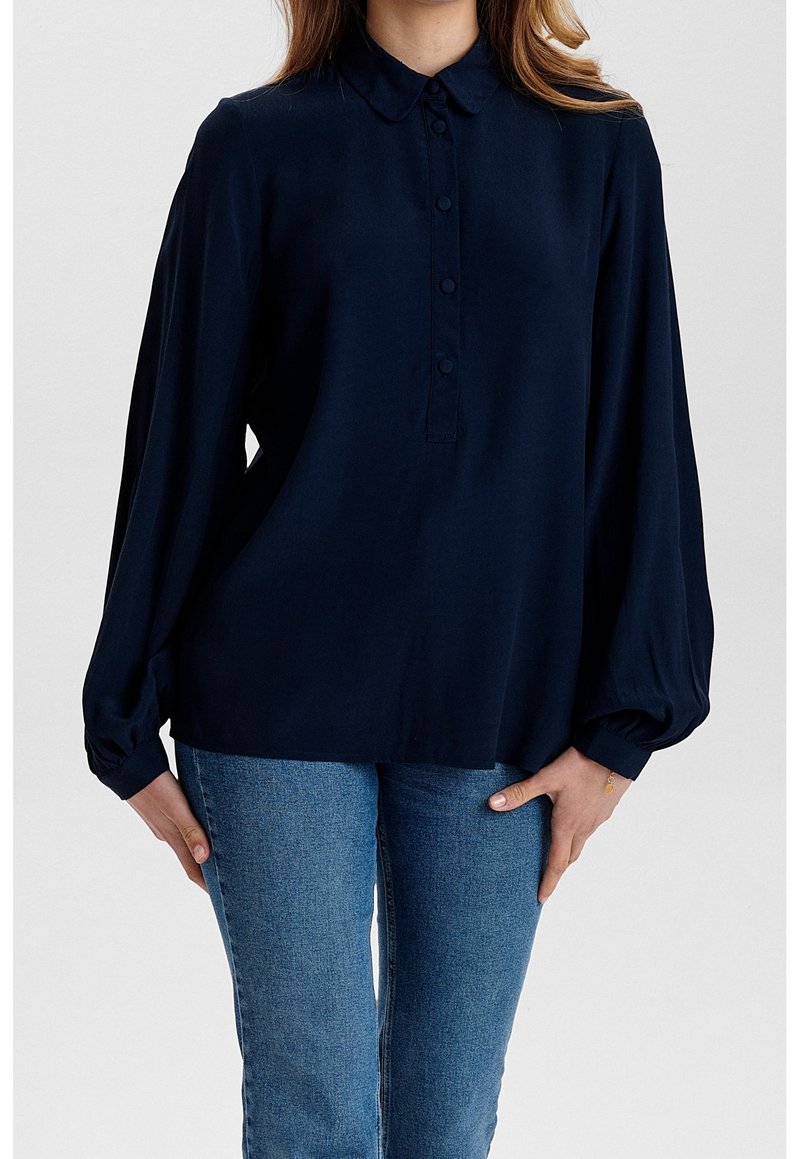 Chemise à manches longues bleu marine avec col, boutonnage à l'avant et coupe ample. Le tissu semble lisse ; les manches sont resserrées aux poignets. Associée à un jean bleu.