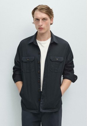 Jonge man met blond haar die een zwart overhemd draagt over een wit T-shirt, handen in de zakken, staand tegen een effen lichte achtergrond.