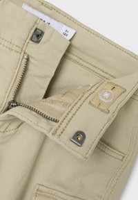 Shorts en tissu beige avec une fermeture éclair et un bouton, dotés de quincaillerie métallique, de détails de couture et d'une étiquette intérieure.